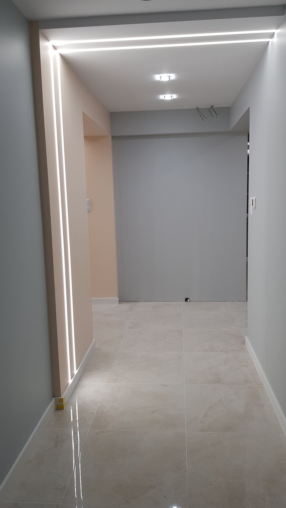 Korytarz z płytkami podłogowymi w odcieniach beżu i szarości, oświetlenie sufitowe LED w formie prostokątnej ramy i pionowych pasów na ścianie, widoczne niedokończone okablowanie na suficie.