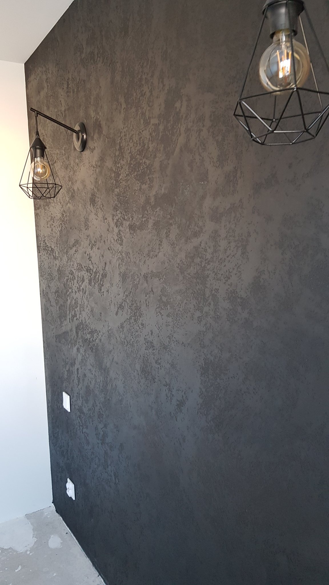 Dekoracyjna, ciemnoszara ściana z efektem betonu, oświetlona dwoma lampami z odsłoniętymi żarówkami w metalowych, geometrycznych kloszach, widoczne gniazdka elektryczne.