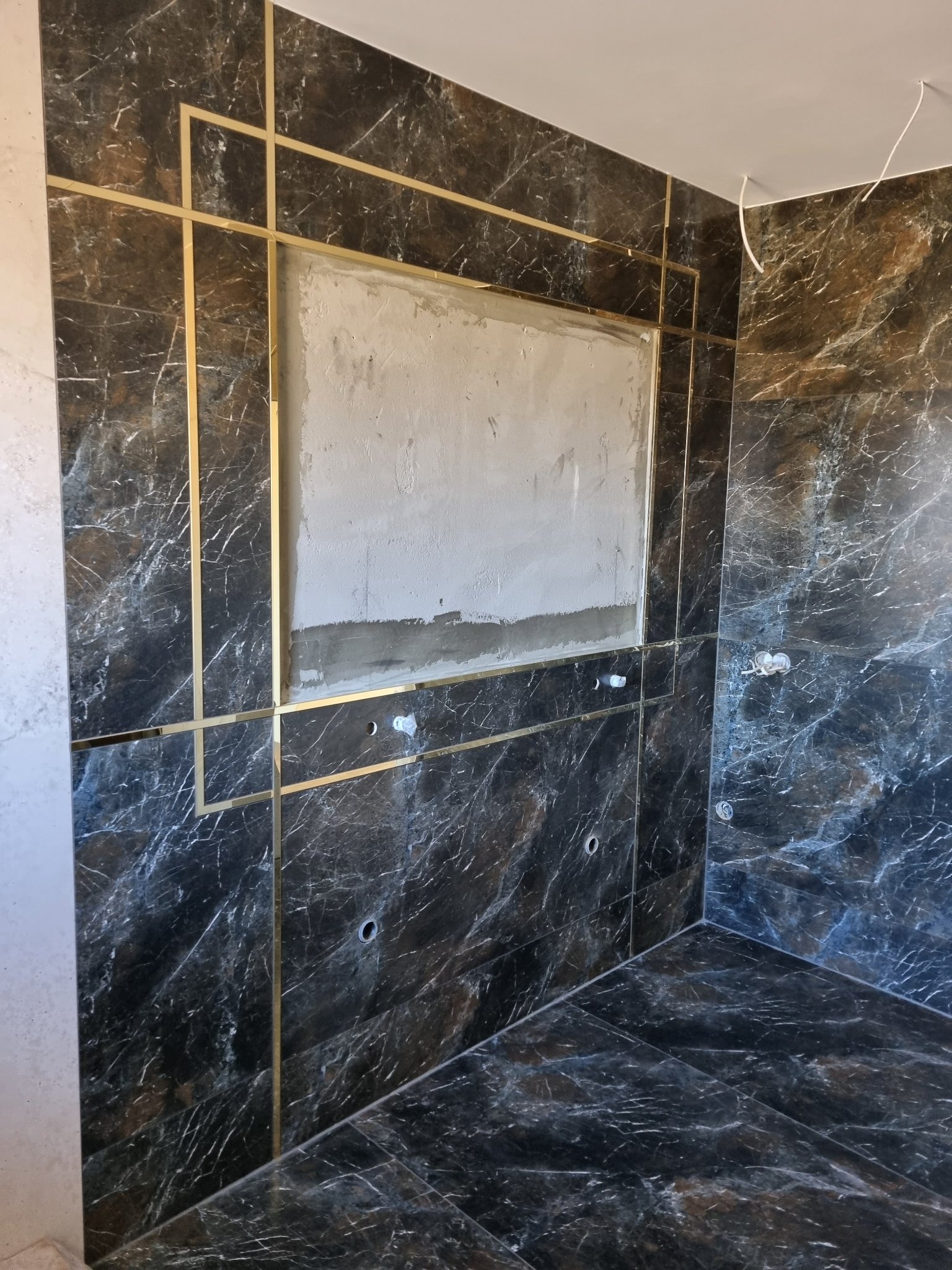 Łazienka w trakcie wykańczania: ciemne marmurowe płytki z złotymi listwami dekoracyjnymi, przygotowane miejsce pod lustro lub szafkę z surową ścianą, widoczne otwory na armaturę.