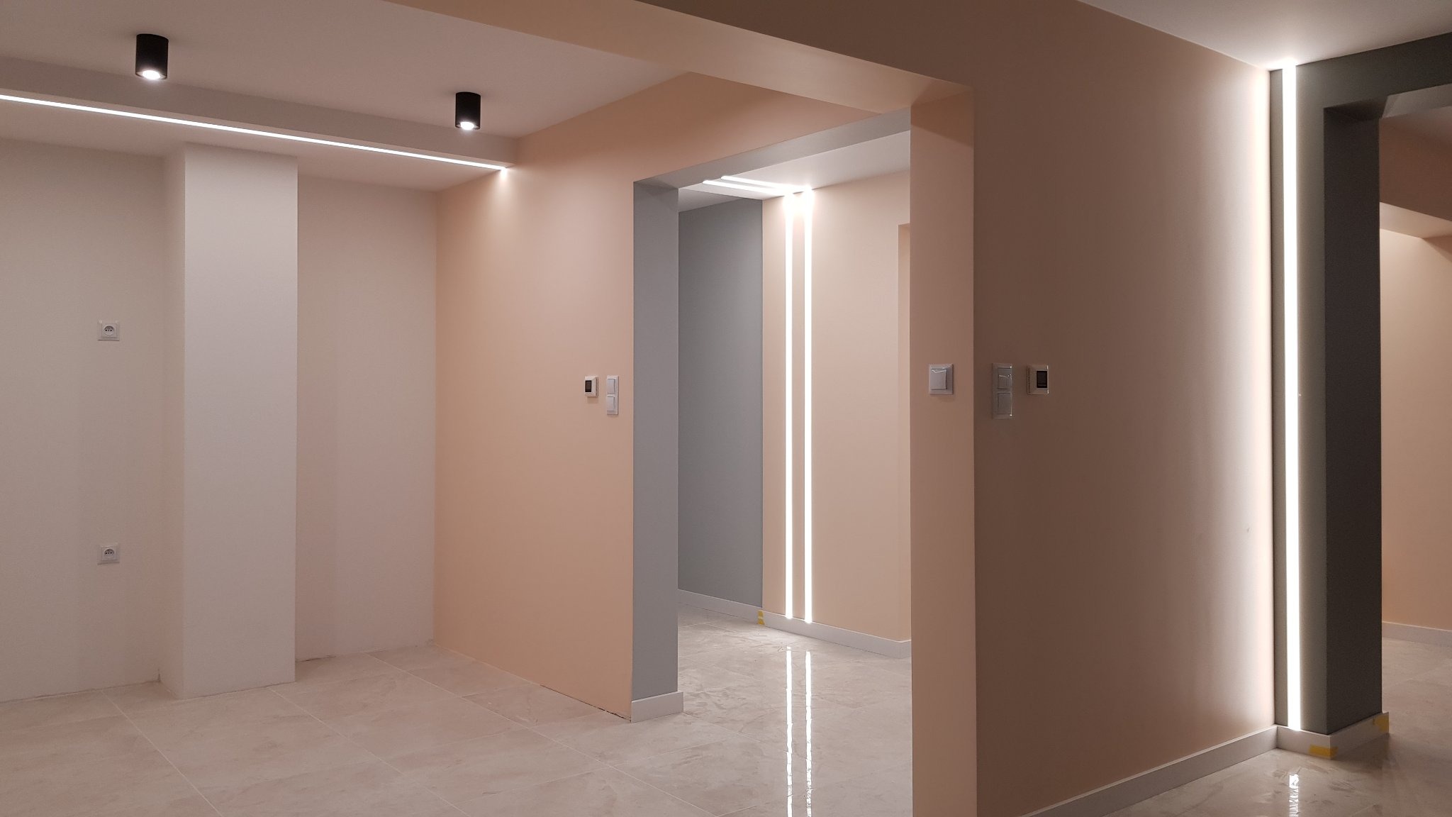 Wykończone wnętrze z jasnymi ścianami w odcieniach beżu i szarości, marmurowa podłoga, oświetlenie liniowe LED na suficie i ścianach, widoczne gniazdka elektryczne i włączniki.