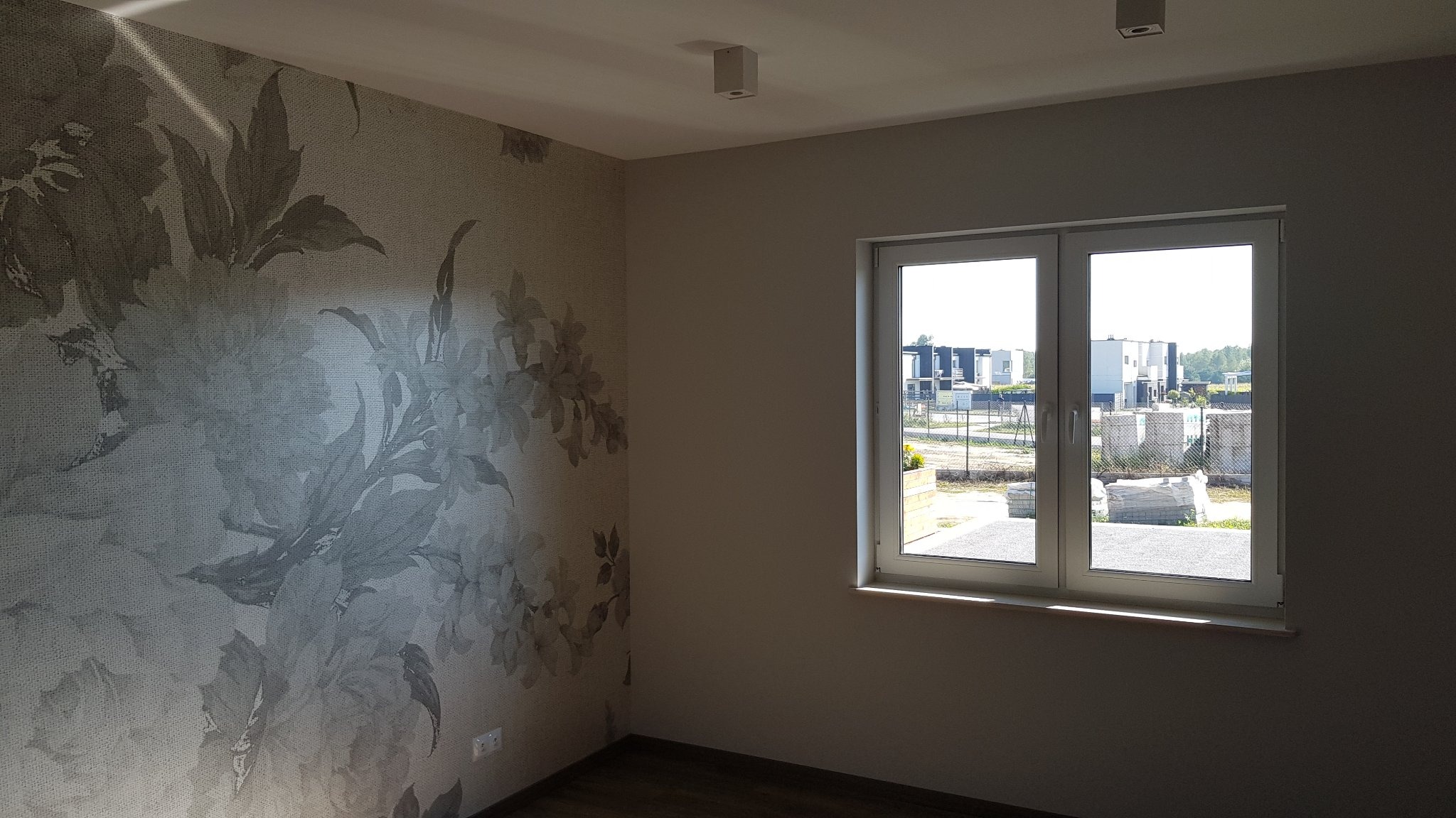 Pokój po remoncie z tapetą w kwiaty na jednej ścianie i oknem z widokiem na osiedle domów.