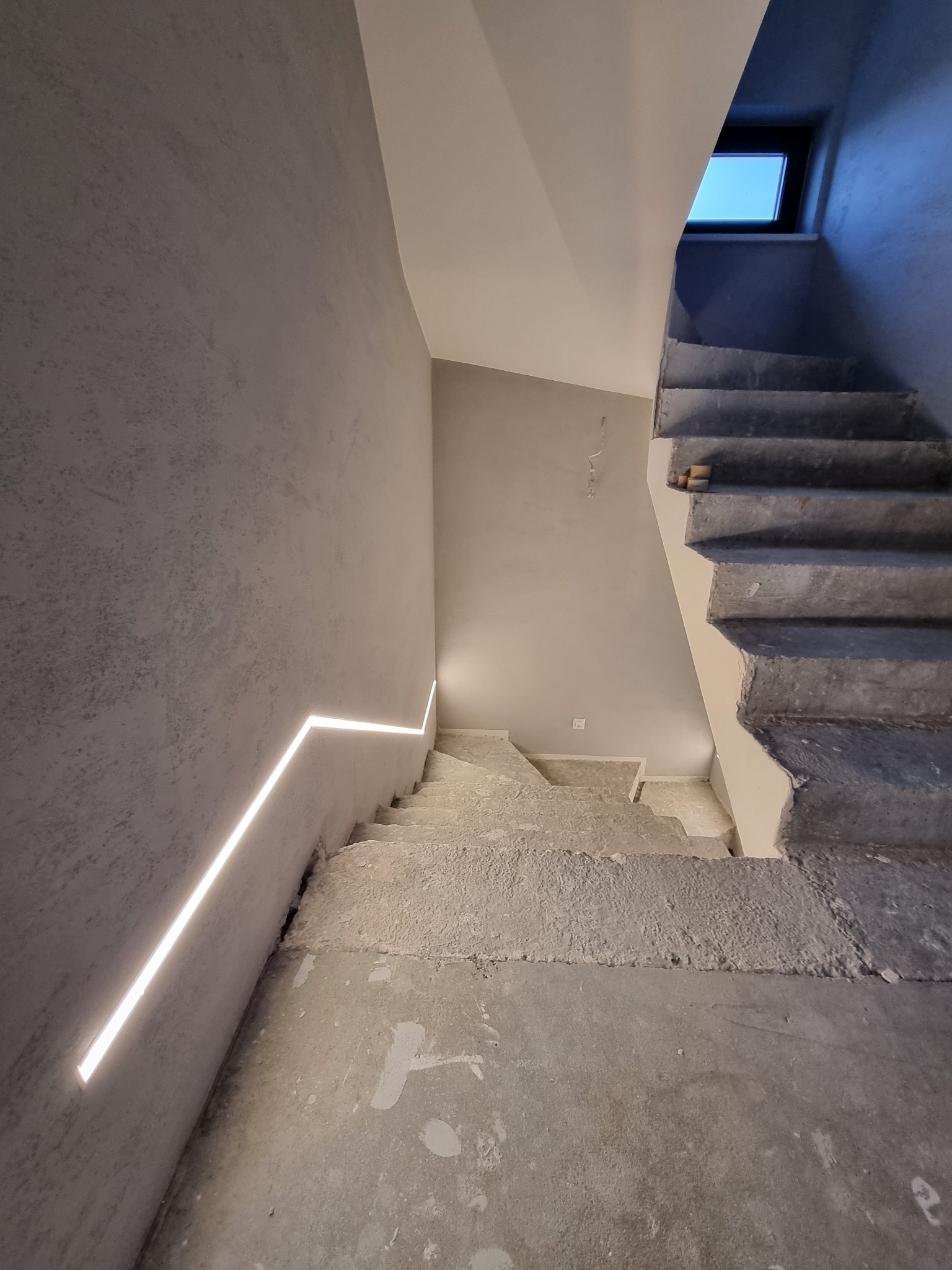 Schody betonowe w trakcie wykańczania, z widocznym oświetleniem LED w ścianie i surowymi ścianami z szarą fakturą, małe okno w górnej części klatki schodowej.