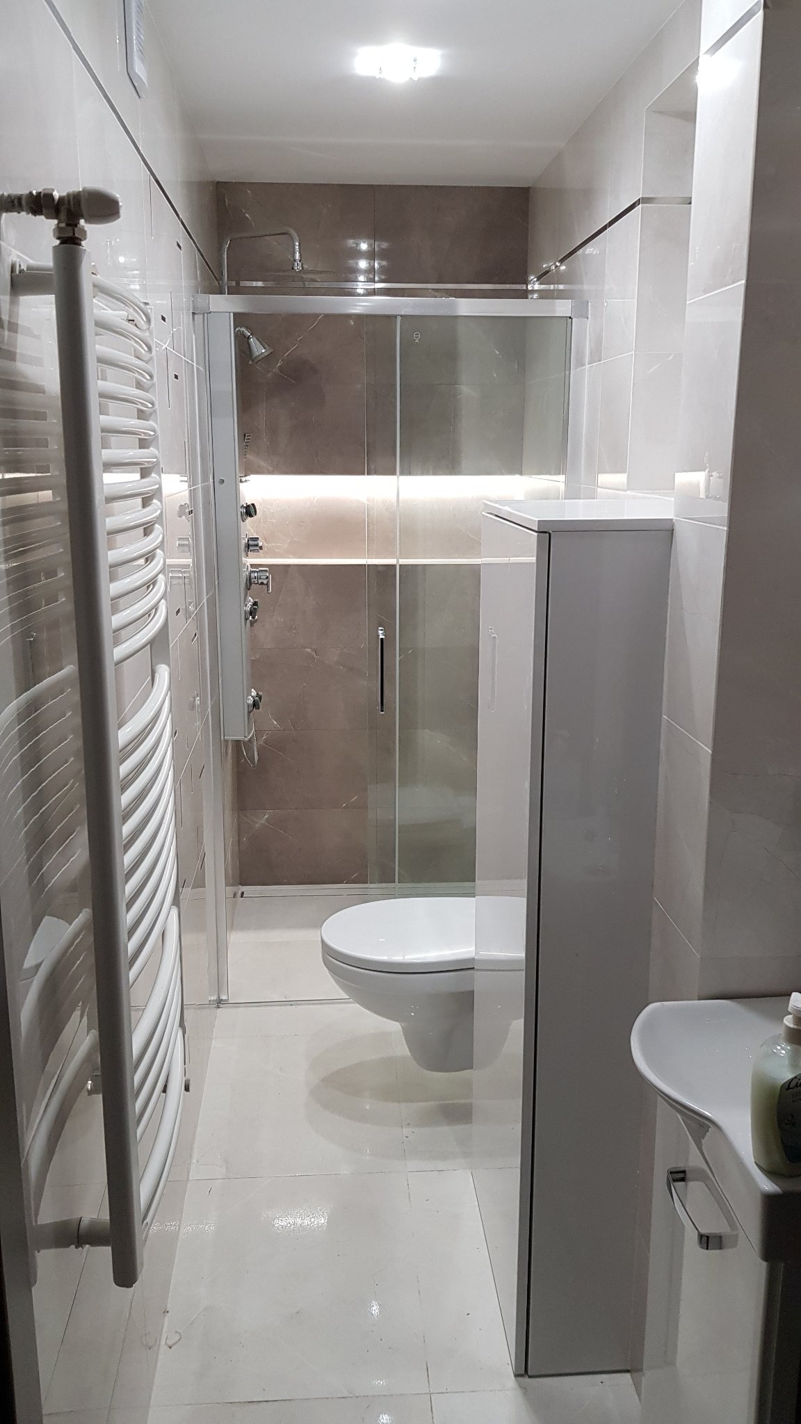 Nowoczesna łazienka z kabiną prysznicową typu walk-in, podświetleniem LED, grzejnikiem drabinkowym i umywalką z szafką, wykończona w jasnych płytkach imitujących marmur.