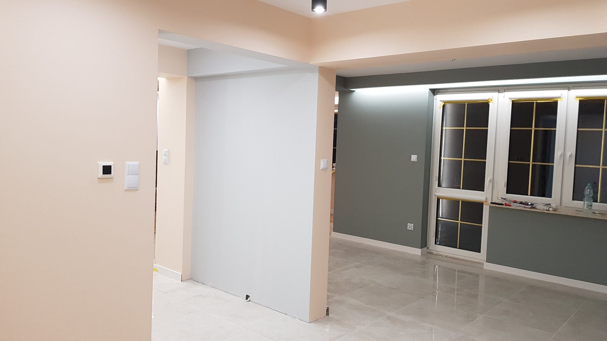 Jasne, przestronne wnętrze po remoncie: kremowe ściany, szare akcenty, połyskująca podłoga z płytek, okna zabezpieczone taśmą malarską. Widoczne gniazdka elektryczne i włączniki.