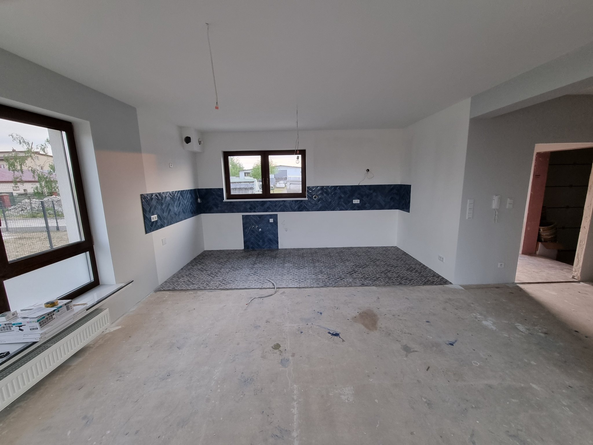 Wnętrze w trakcie wykończenia: częściowo ułożona podłoga z wzorzystych płytek, pas ciemnoniebieskich płytek na ścianie, surowy beton na pozostałej powierzchni podłogi, przewody wystające z sufitu.