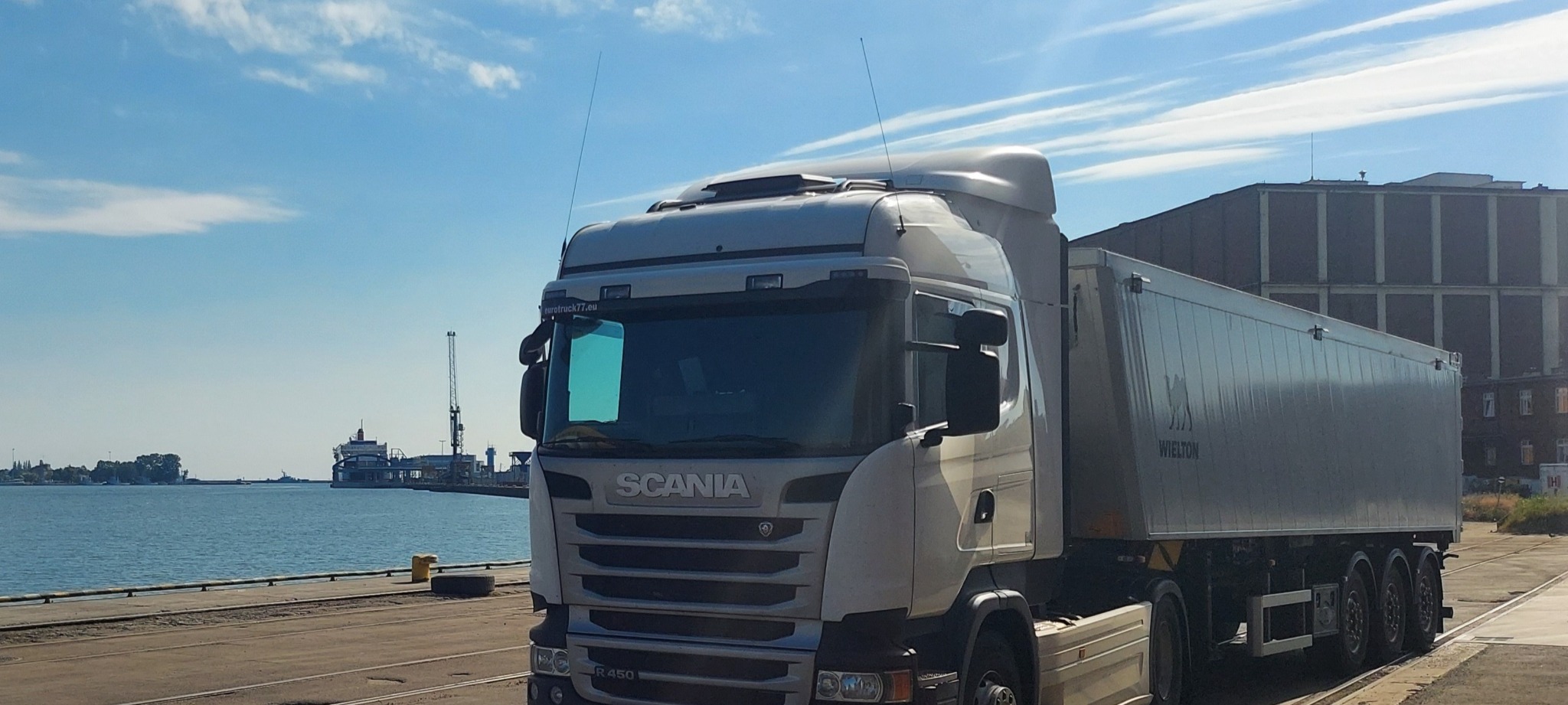 Ciężarówka Scania z naczepą Wielton na tle portu i błękitnego nieba. Widoczny dźwig portowy i budynek magazynowy.