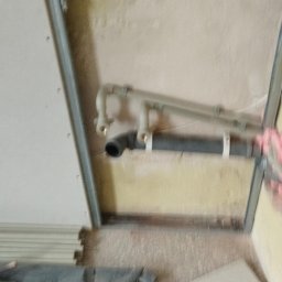 BORKOWSKI DAMIAN - Wnętrze łazienki w trakcie remontu: widoczne rury kanalizacyjne przymocowane do ściany, fragment płyty karton gips z metalowym profilem oraz fragment podłogi z płytkami.