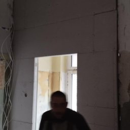 BORKOWSKI DAMIAN - Montaż płyt kartonowo-gipsowych na ścianie z widoczną konstrukcją metalową sufitu i instalacją elektryczną; w tle widoczne drzwi i fragment pomalowanej ściany.