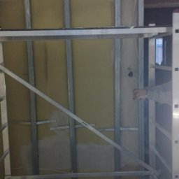 BORKOWSKI DAMIAN - Metalowy stelaż pod płytę karton-gips zamontowany na żółtej ścianie, widoczna drabina Krause i fragment sylwetki pracownika