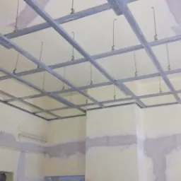 Metalowa konstrukcja sufitu podwieszanego z płyt kartonowo-gipsowych, widoczne profile i zawiesia, ściany w tle z widocznymi śladami szpachlowania.