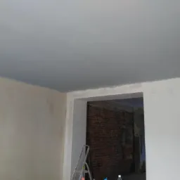 Świeżo pomalowany sufit na szaro w pokoju, widoczna drabina i fragment ceglanej ściany w przejściu.