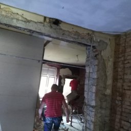 BORKOWSKI DAMIAN - Wnętrze w trakcie remontu: płyta kartonowo-gipsowa oparta o ścianę, odsłonięta cegła, widoczni pracownicy w trakcie pracy.