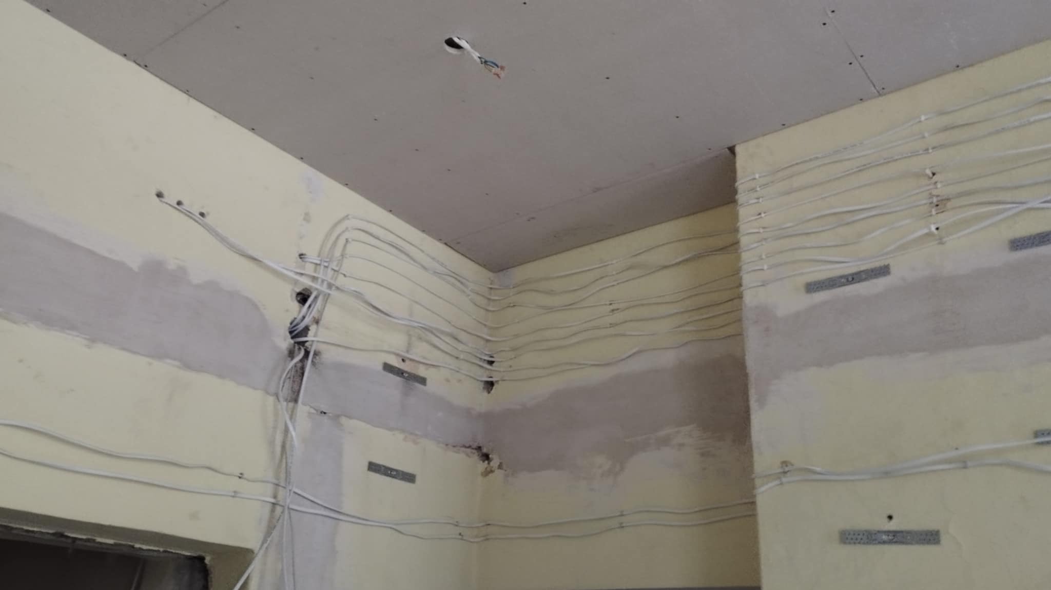 Ściana w trakcie remontu z widocznymi przewodami elektrycznymi ułożonymi poziomo i przymocowanymi do ściany za pomocą metalowych uchwytów, ściana częściowo zagruntowana, sufit z płyt...