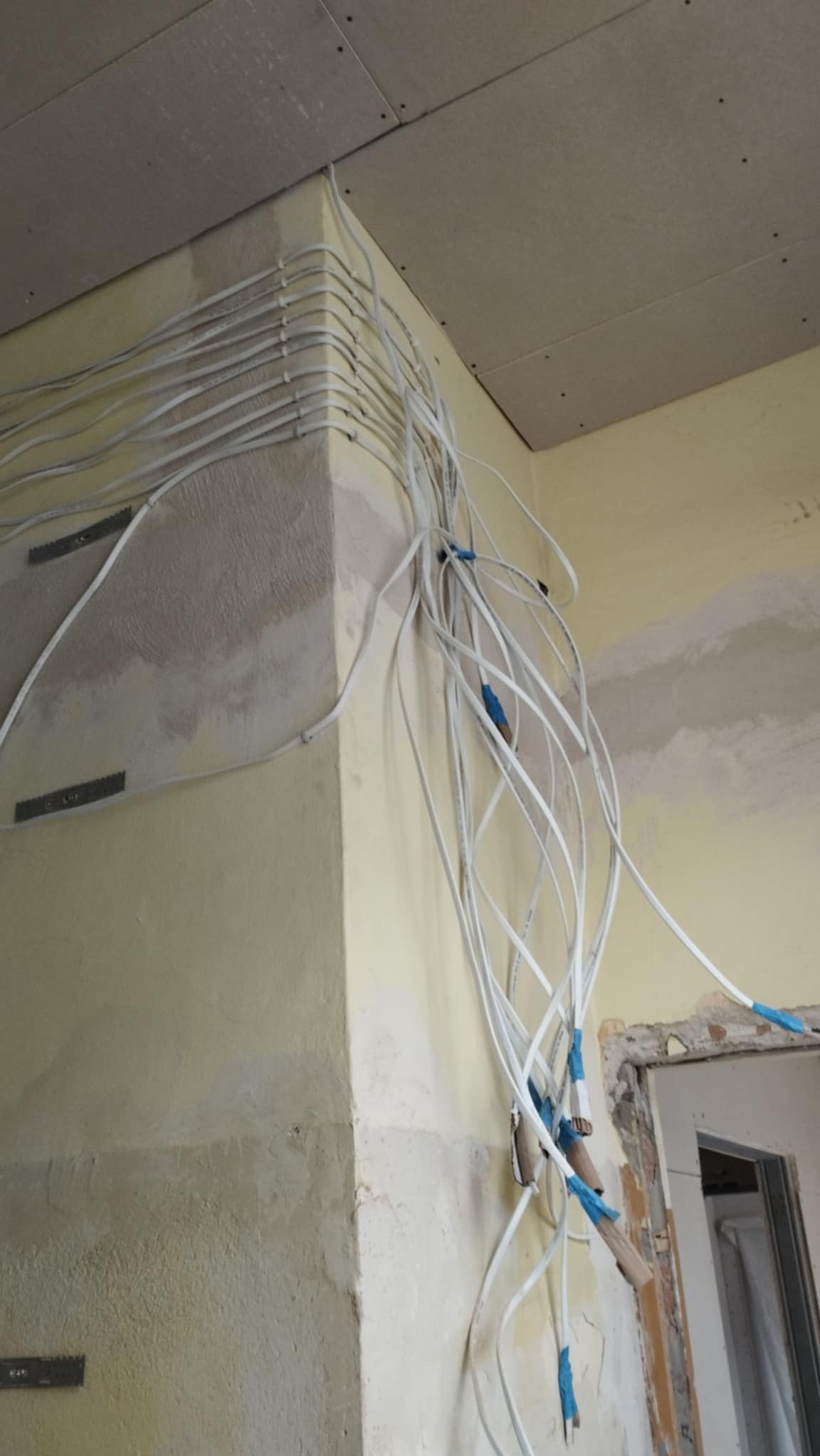 Ściana narożna z licznymi przewodami elektrycznymi ułożonymi wzdłuż krawędzi, zabezpieczonymi niebieską taśmą, widoczne ślady po szpachlowaniu i płyta karton gips na suficie, fragment ościeżnicy...