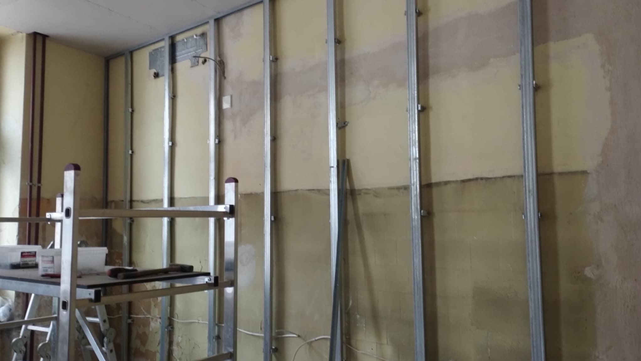 Montaż metalowej konstrukcji szkieletowej do płyt kartonowo-gipsowych na nierównej ścianie w trakcie remontu, widoczny aluminiowy ruszt i narzędzia.