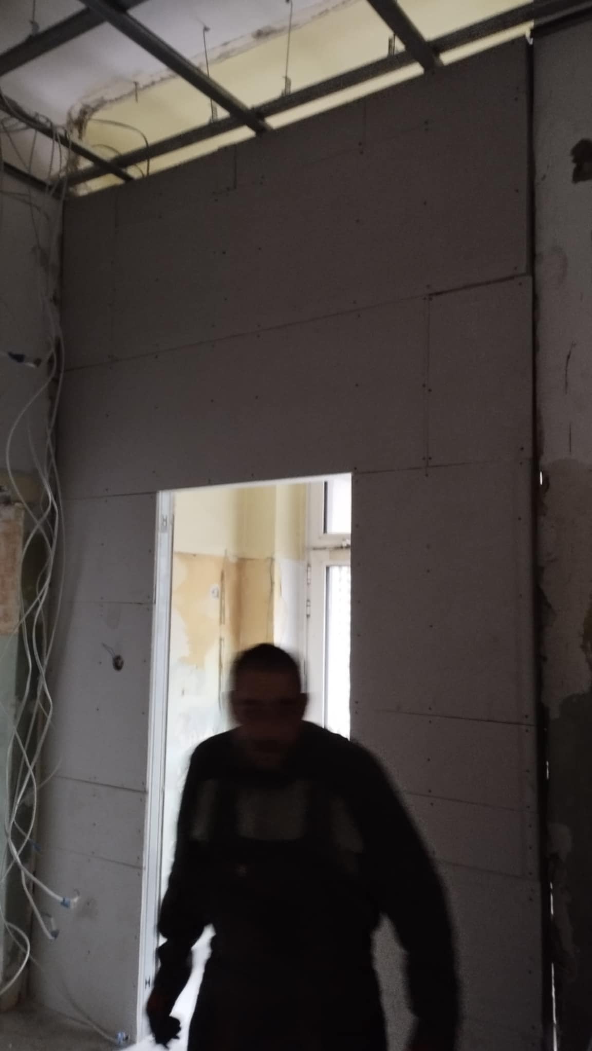 Montaż płyt kartonowo-gipsowych na ścianie z widoczną konstrukcją metalową sufitu i instalacją elektryczną; w tle widoczne drzwi i fragment pomalowanej ściany.