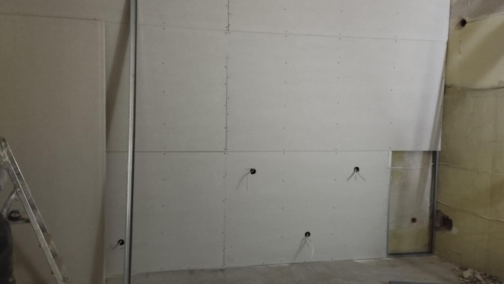 Montaż płyt kartonowo-gipsowych na ścianie z widocznymi otworami na instalacje elektryczne i fragmentem starej, nieotynkowanej ściany w tle.