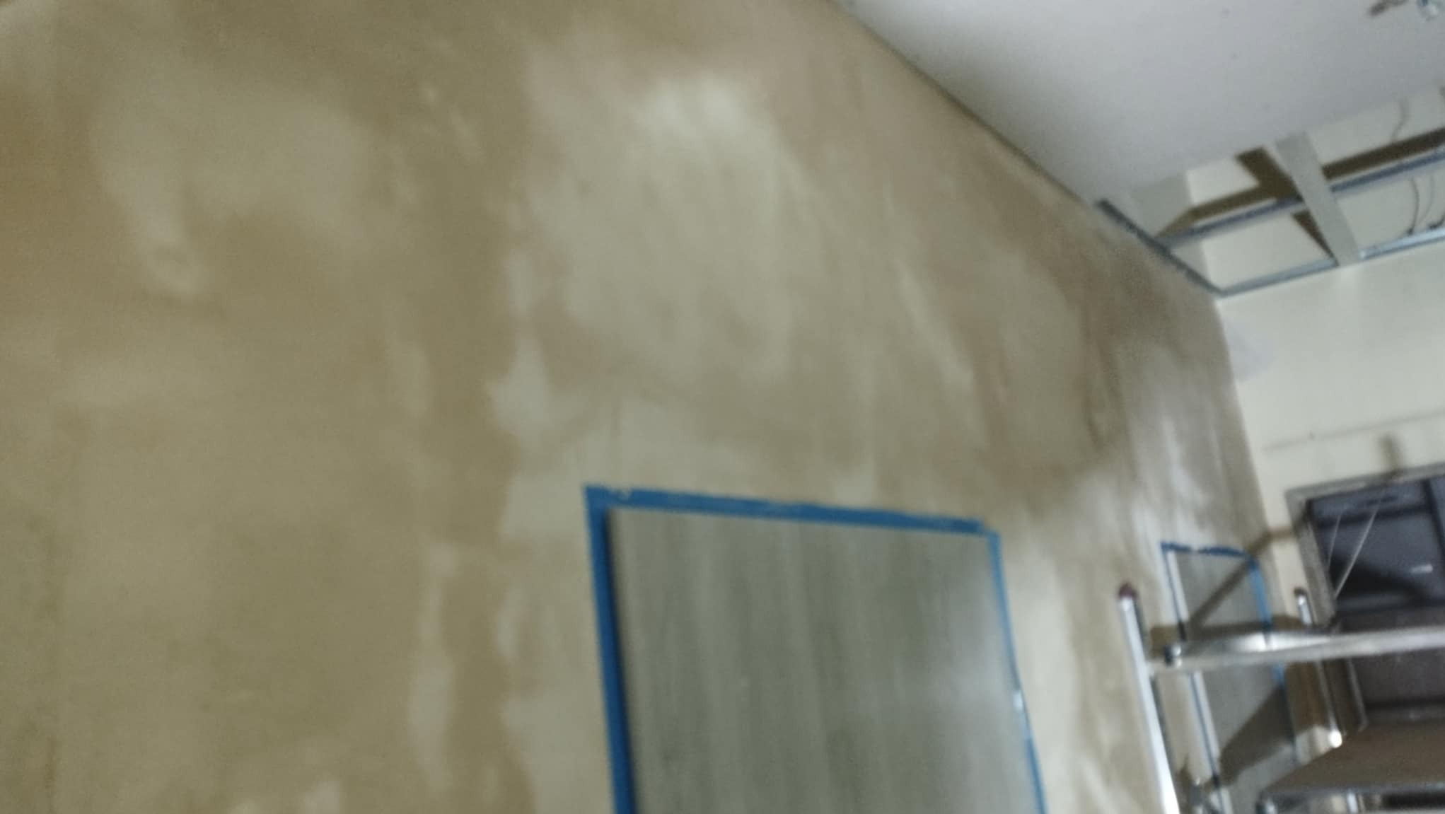 Ściana po nałożeniu pierwszej warstwy gładzi, z widocznym zabezpieczeniem folią malarską framugi drzwiowej i rusztowaniem w tle.