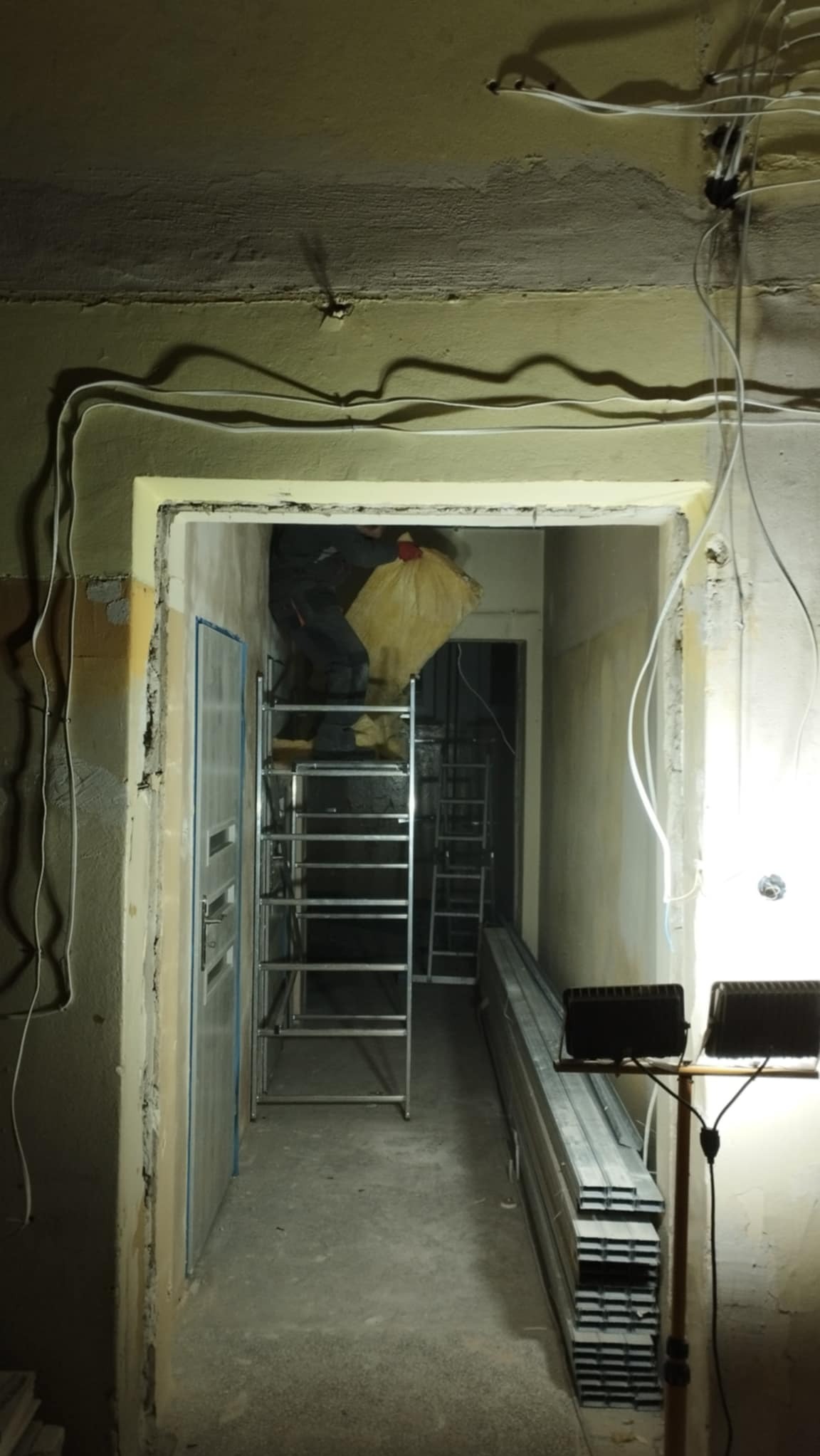 Wnętrze w trakcie remontu: widoczne przewody elektryczne na suficie, metalowe profile ułożone wzdłuż ściany, rozstawiony ruszt aluminiowy, pracownik w kombinezonie z workiem śmieci.