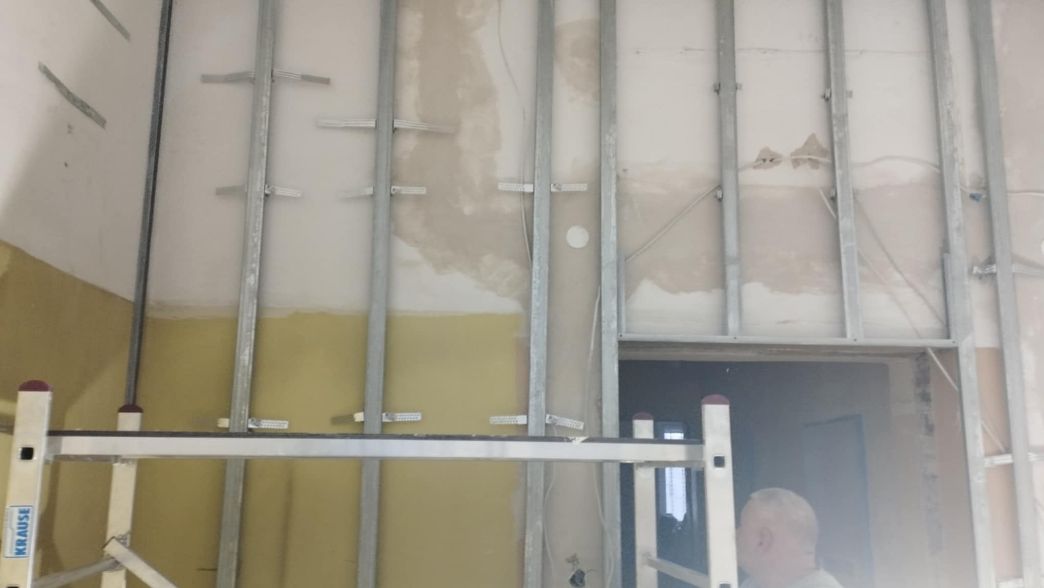 Montaż metalowego stelażu pod płyty karton gips na ścianie, widoczne fragmenty instalacji elektrycznej i drabina Krause w pomieszczeniu w trakcie remontu.