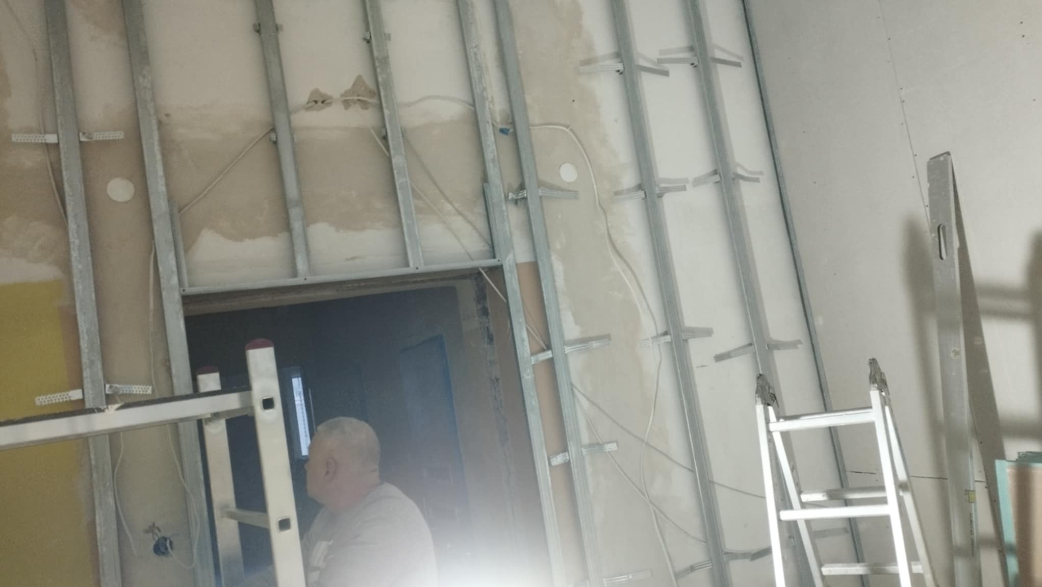 Montaż metalowego stelażu do płyt kartonowo-gipsowych na ścianie, widoczne przewody elektryczne i drabina, mężczyzna w tle.