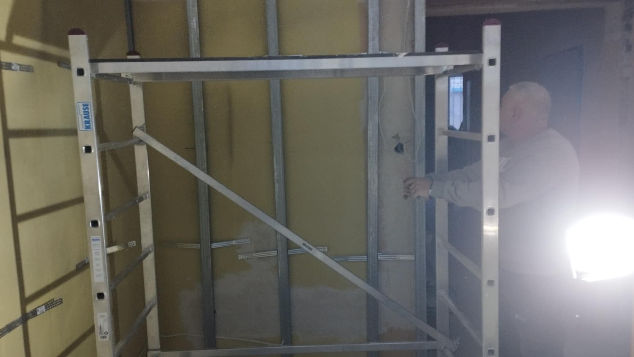 Metalowy stelaż pod płytę karton-gips zamontowany na żółtej ścianie, widoczna drabina Krause i fragment sylwetki pracownika