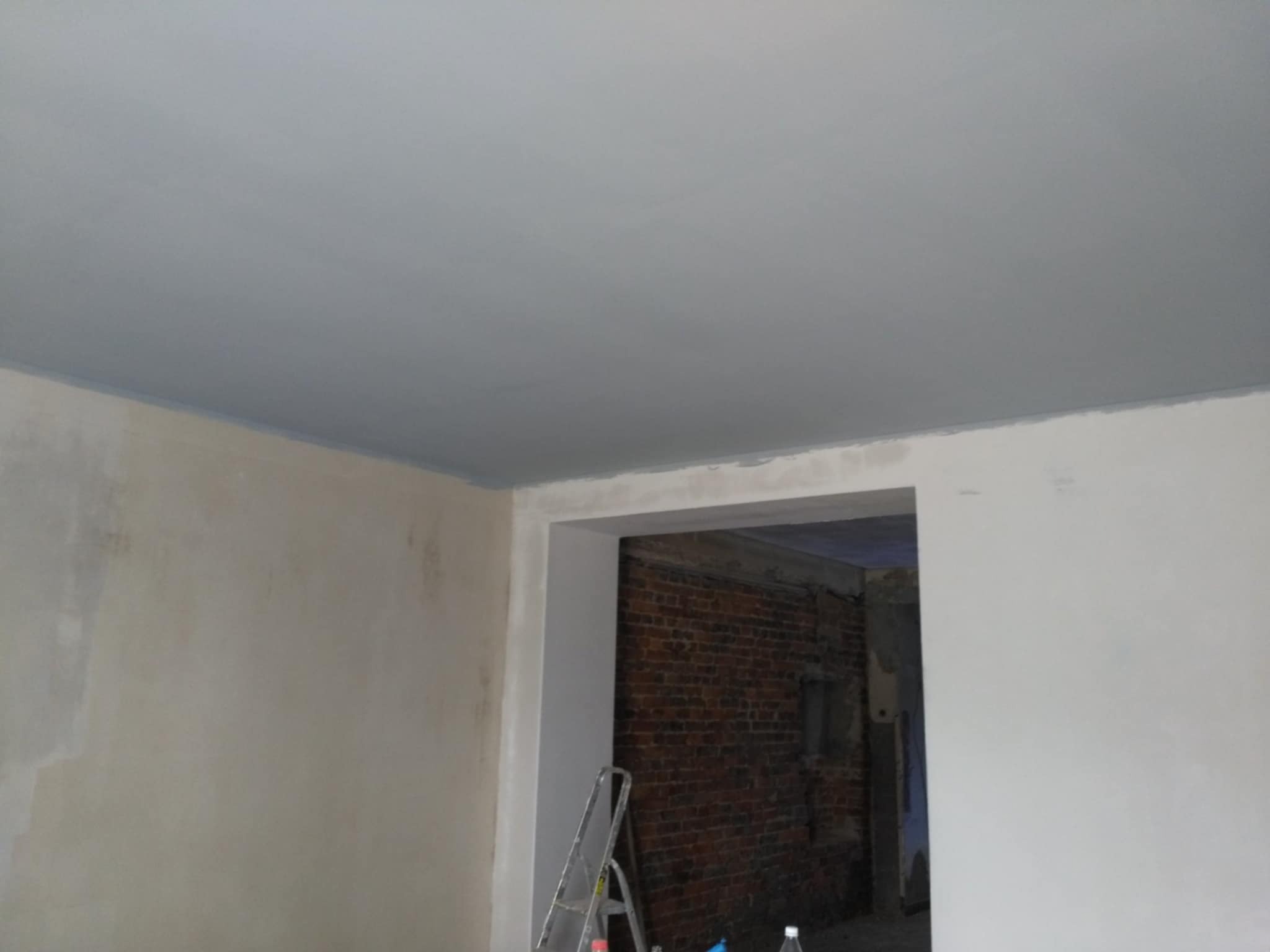 Świeżo pomalowany sufit na szaro w pokoju, widoczna drabina i fragment ceglanej ściany w przejściu.