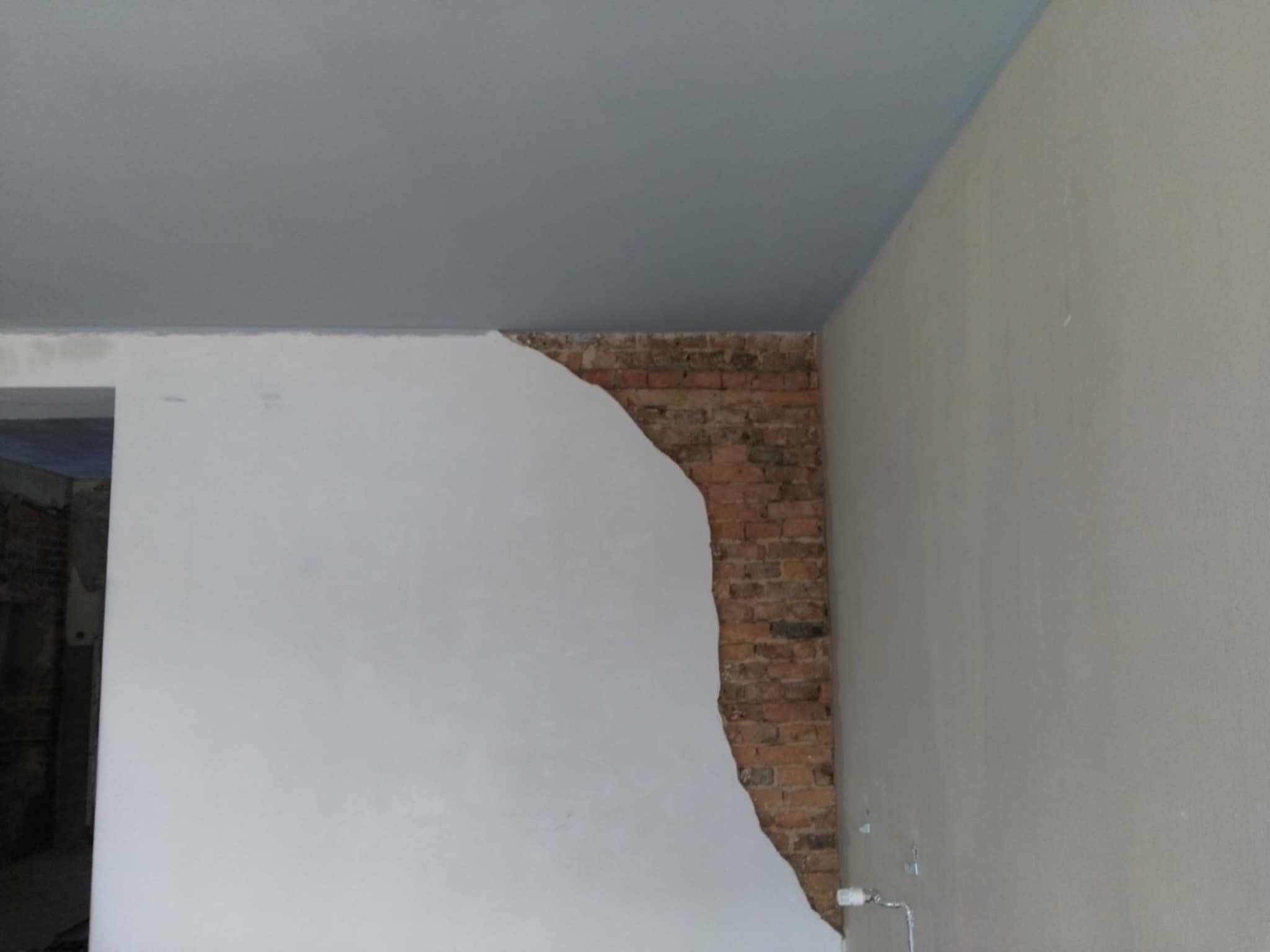 Fragment ściany z odsłoniętą czerwoną cegłą, częściowo pokrytej świeżo nałożonym białym tynkiem, z pomalowanym na szaro sufitem w rogu pomieszczenia, ukazujący etap prac wykończeniowych.