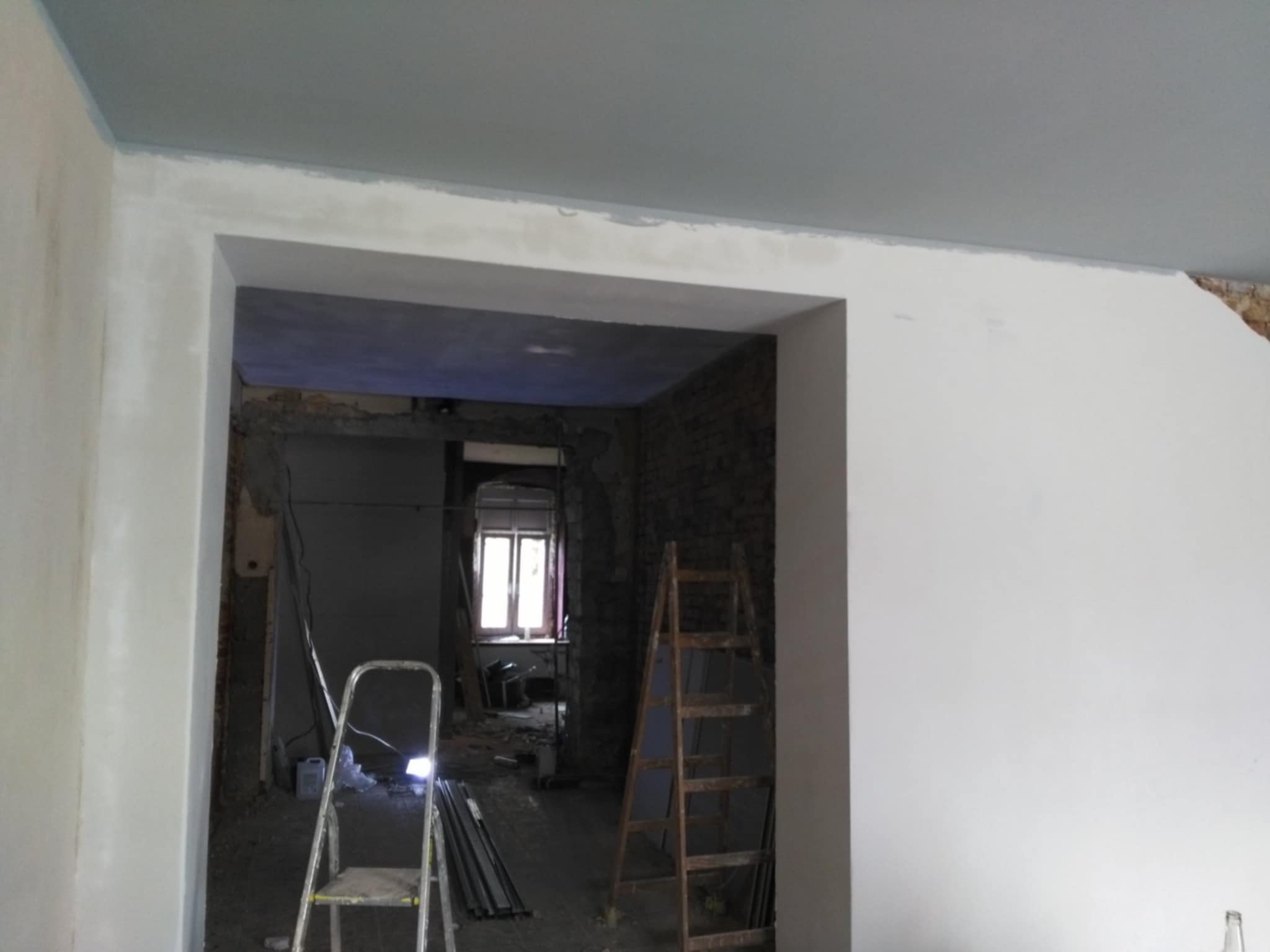 Wnętrze w trakcie remontu: pomalowany sufit na szaro, ściany częściowo zagruntowane na biało, widoczne dwie drabiny i fragment okna w głębi pomieszczenia.