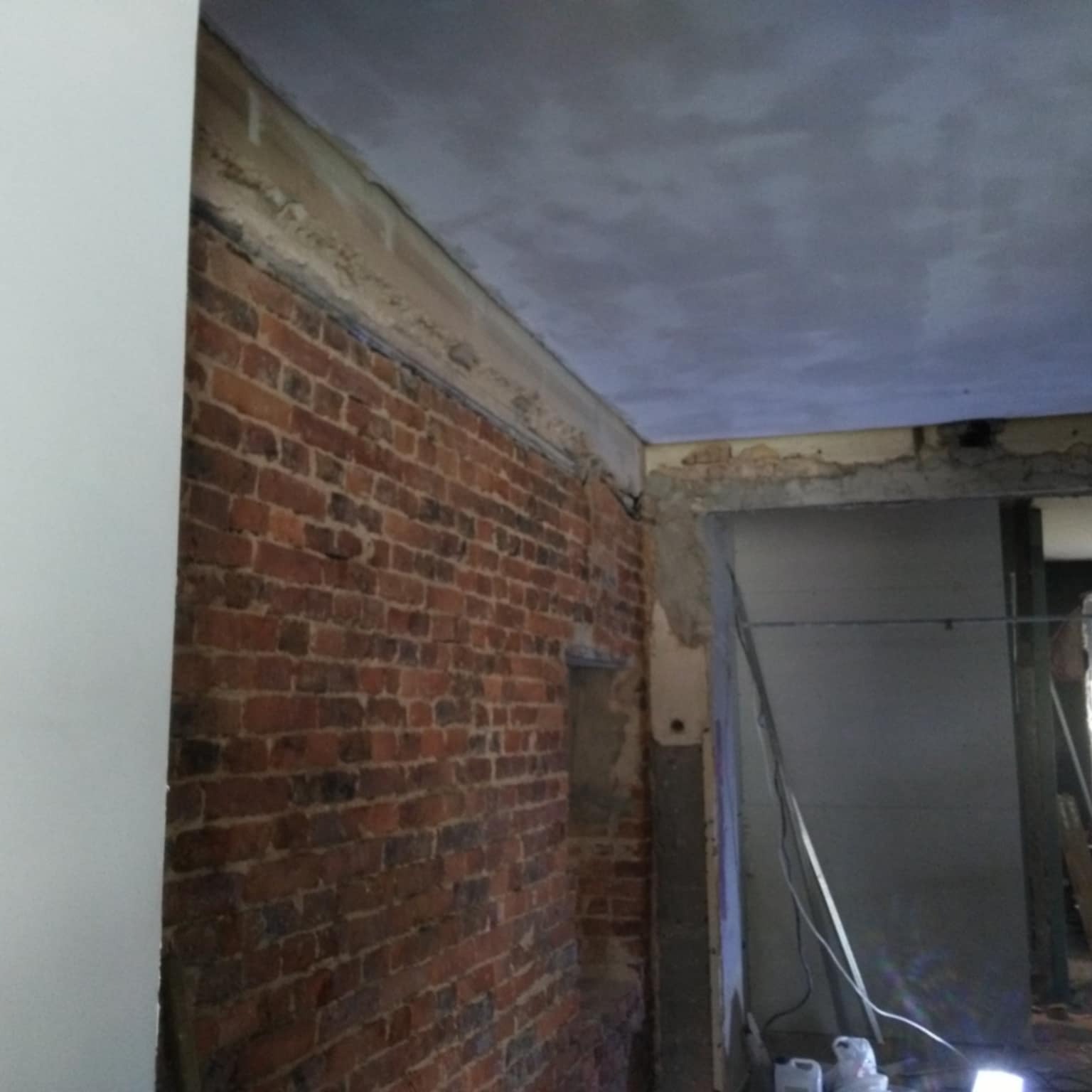 Częściowo wykończone wnętrze z odsłoniętą ceglaną ścianą, świeżo zagruntowanym sufitem i przygotowanym otworem drzwiowym.