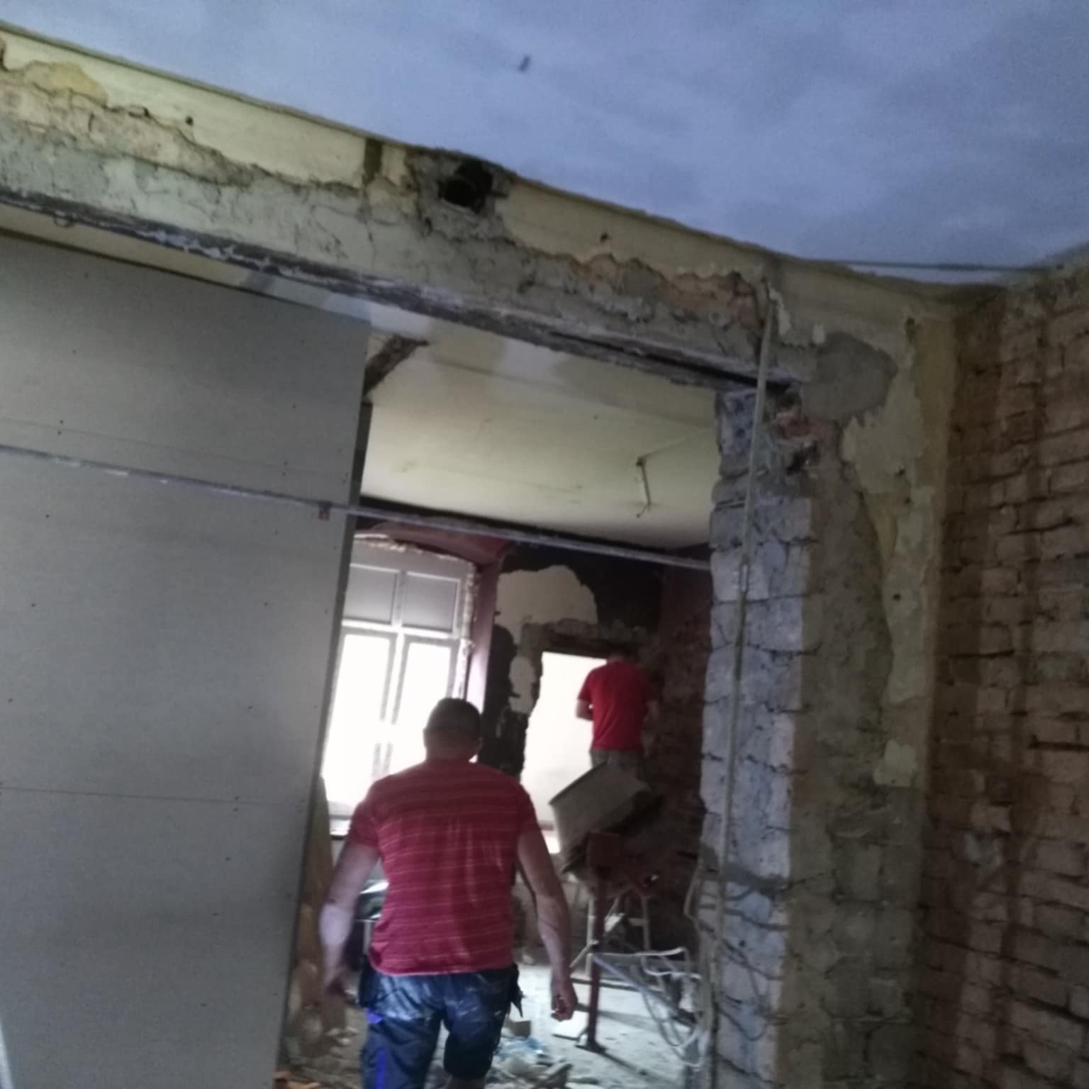 Wnętrze w trakcie remontu: płyta kartonowo-gipsowa oparta o ścianę, odsłonięta cegła, widoczni pracownicy w trakcie pracy.