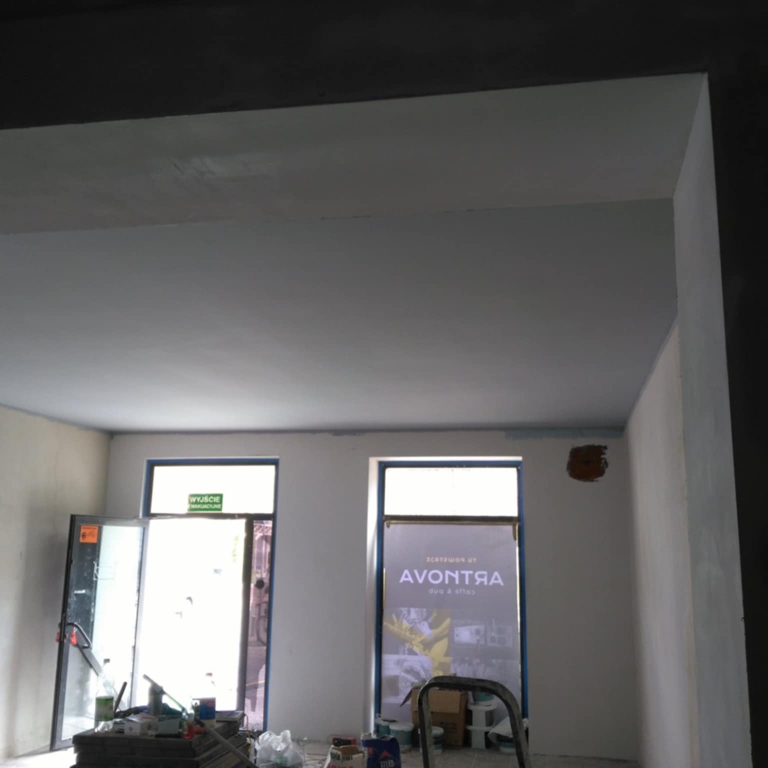 Wnętrze w trakcie malowania: świeżo pomalowany sufit, częściowo pomalowane ściany z widocznymi poprawkami i otwartymi drzwiami ewakuacyjnymi, narzędzia malarskie i drabina w tle.