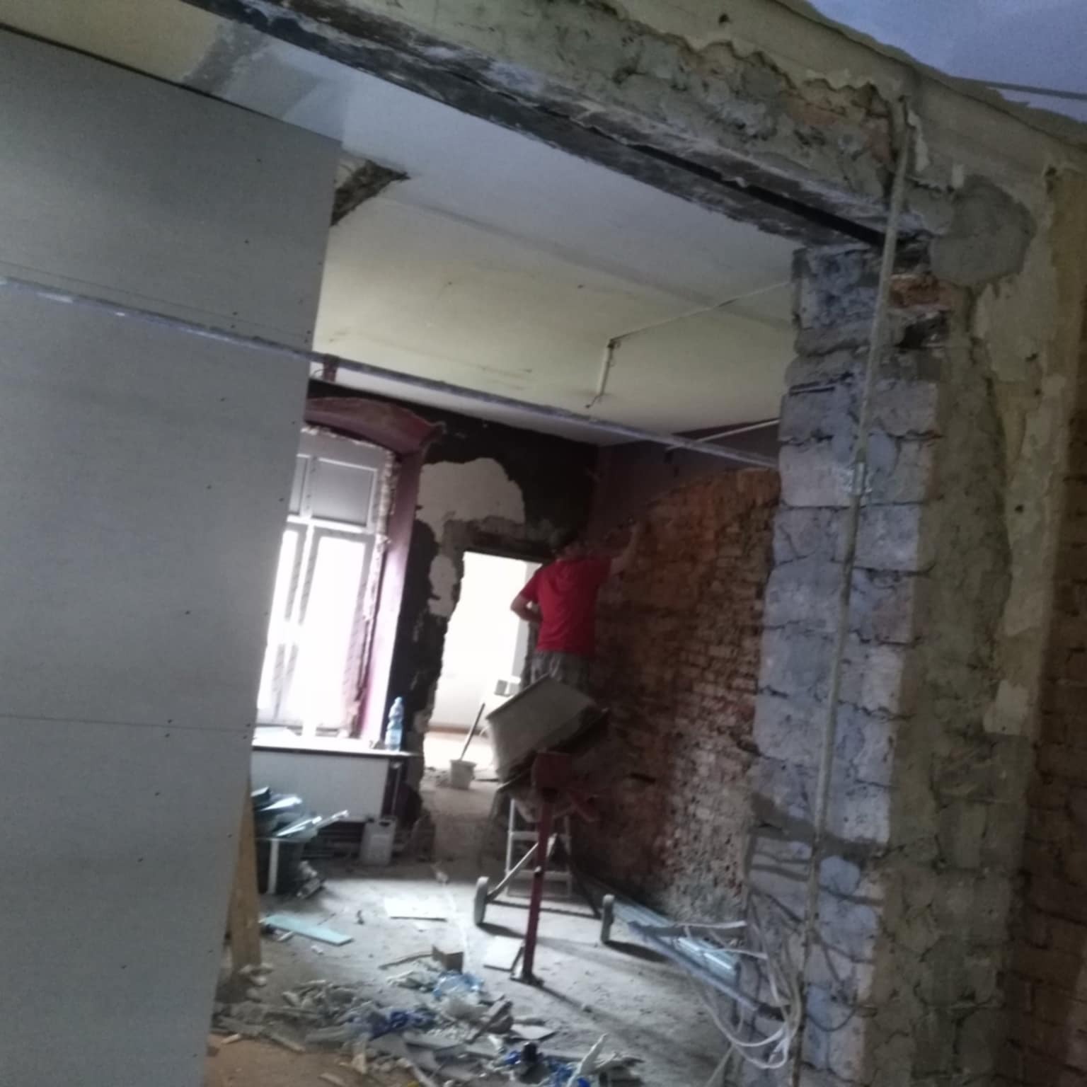 Prace remontowe w trakcie w Mysłowicach: montaż płyty karton gips na ścianie, widoczny fragment odsłoniętej cegły i pracownik w czerwonej koszulce.