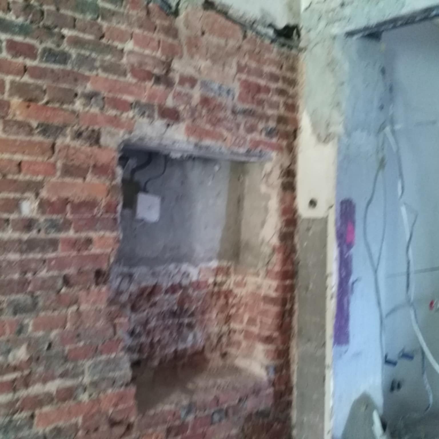 Ceglana ściana z wyciętym otworem, widoczne przewody elektryczne i fragment ściany z naniesionymi bazgrołami w trakcie prac remontowych.