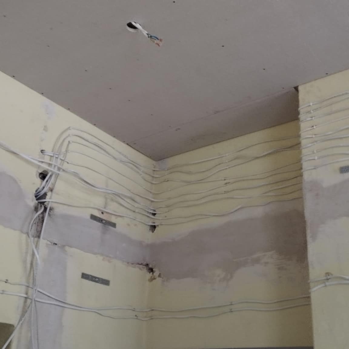 Ściana w trakcie remontu z widocznymi przewodami elektrycznymi ułożonymi poziomo, częściowo zakryta płytami kartonowo-gipsowymi, sufit z widocznym otworem na przewody.