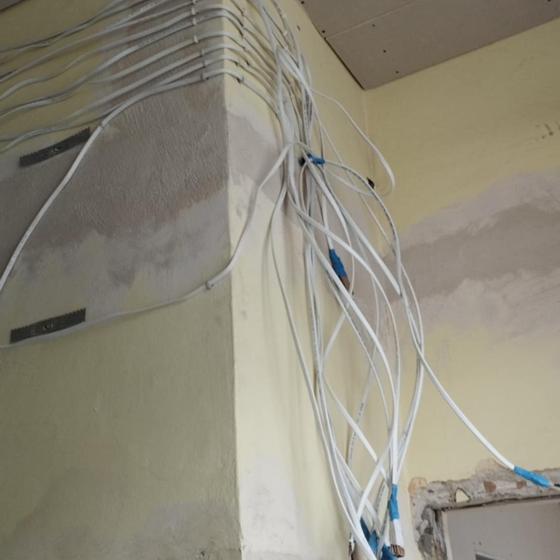 Kąt pokoju z widocznymi przewodami elektrycznymi przymocowanymi do ściany i zwisającymi luźno, ściany w trakcie przygotowania do malowania, widoczna płyta kartonowo-gipsowa na suficie.