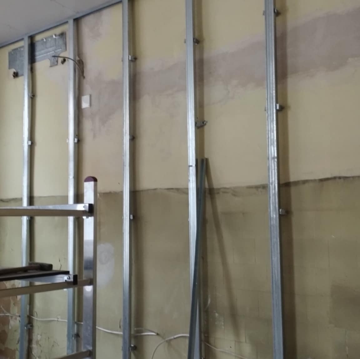 Stelaż z profili metalowych przymocowany do ściany przygotowanej pod montaż płyt gipsowo-kartonowych, widoczne ślady po wcześniejszych pracach na ścianie i fragment rusztowania.