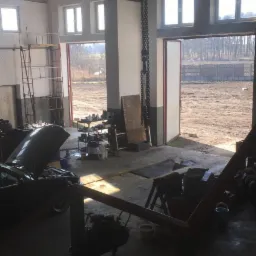 Wnętrze warsztatu samochodowego z podniesioną maską samochodu, widoczne regały z częściami, narzędzia oraz dźwig łańcuchowy pod sufitem, w tle otwarte wrota z widokiem na plac.