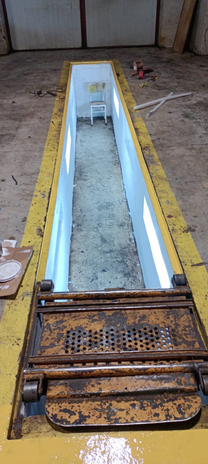 Pomalowany na biało kanał samochodowy z żółtymi krawędziami i metalową kratą ochronną, w głębi widoczne krzesło.