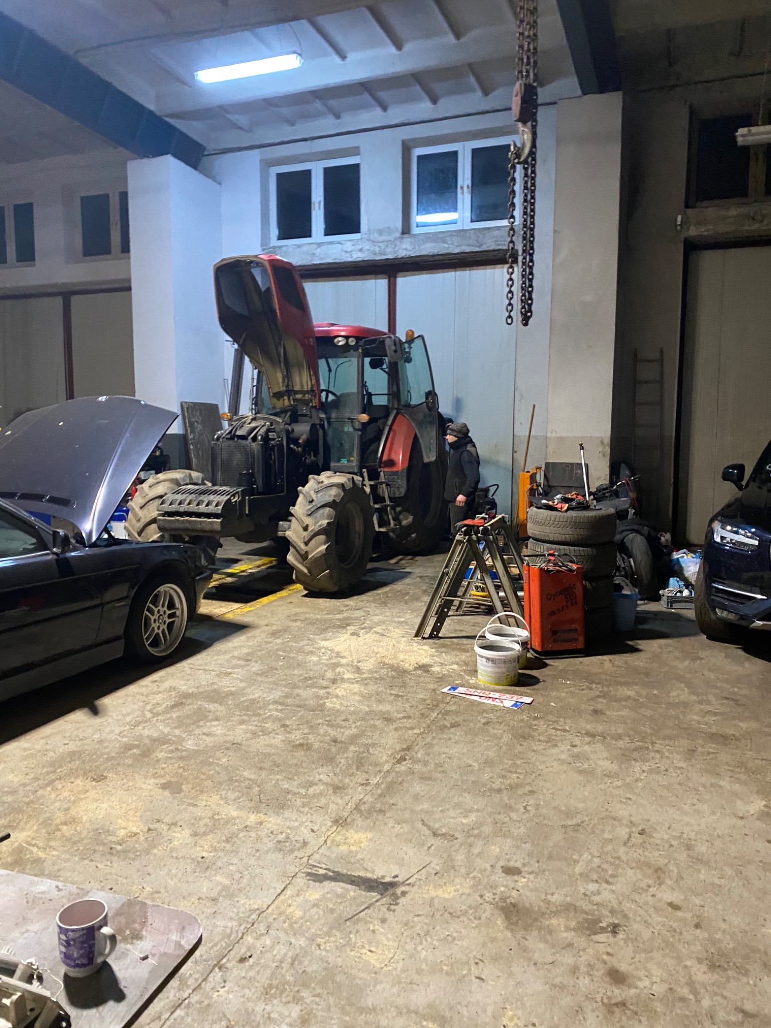 Warsztatowy garaż z podniesioną maską samochodu BMW, czerwonym traktorem z otwartą maską, stosem opon i narzędziami pod łańcuchem z hakiem pod sufitem.