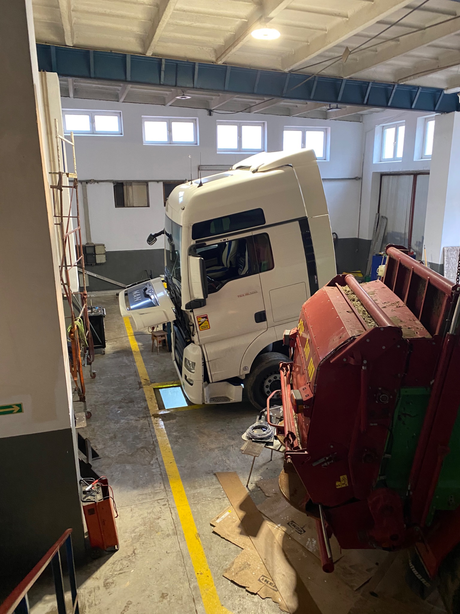 Biała ciężarówka MAN z podniesioną kabiną w warsztacie, obok czerwona maszyna rolnicza, widoczny akumulator na wózku, żółta linia na podłodze.