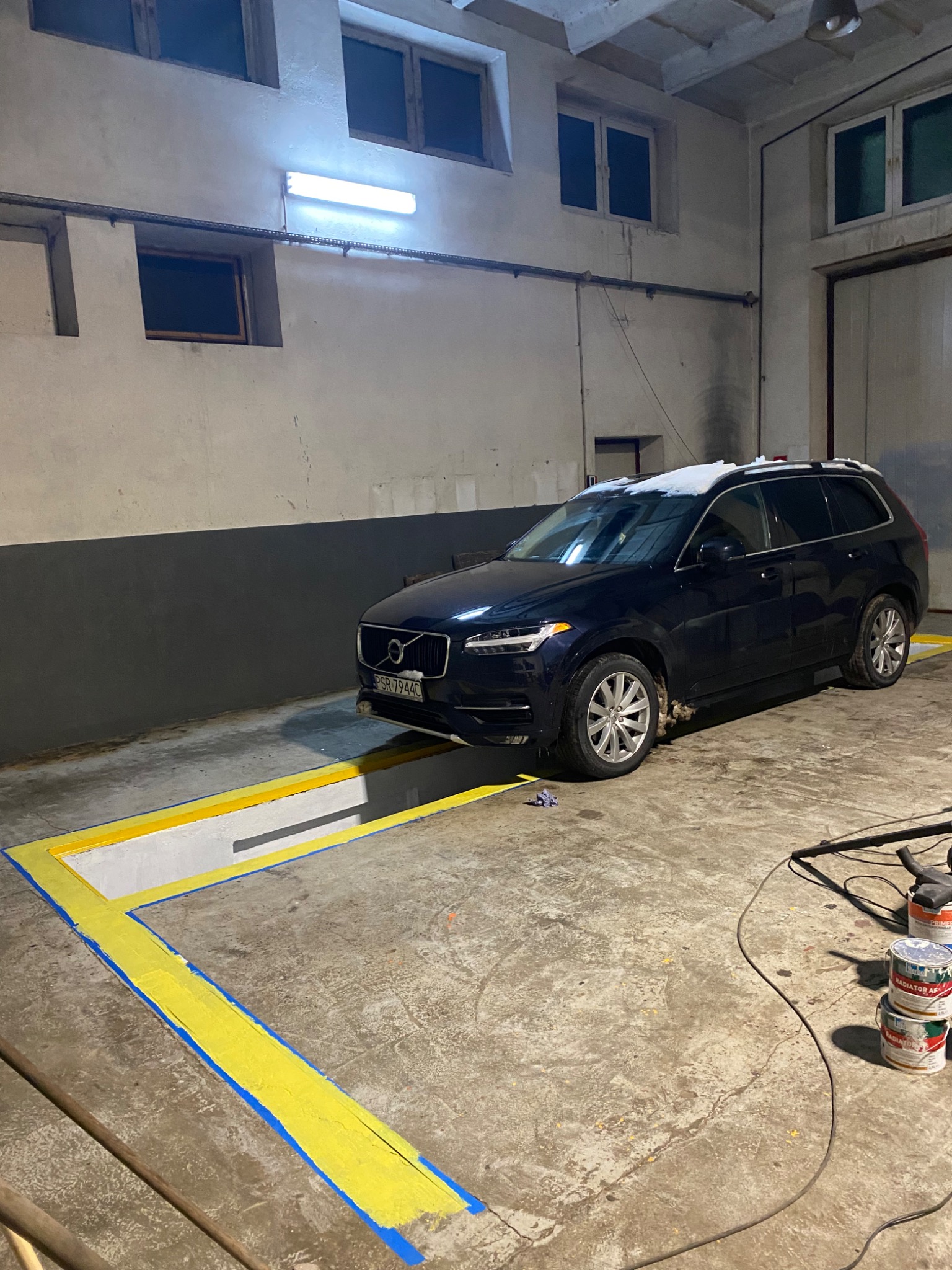 Niebieski samochód osobowy Volvo XC90 z tablicą rejestracyjną PSR 7944C stoi w warsztacie nad kanałem naprawczym oznaczonym żółto-niebieską farbą, widoczne puszki z farbą i przewód odkurzacza.