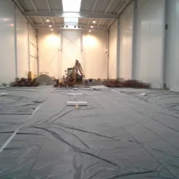 Wnętrze hali w trakcie budowy, podłoże wyłożone folią, w tle koparka i materiały budowlane.