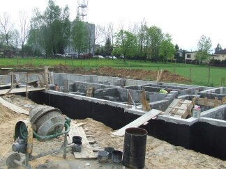 Fundamenty domu jednorodzinnego w trakcie budowy, widoczne szalunki, izolacja przeciwwilgociowa, betonowa podstawa i betoniarka na pierwszym planie.