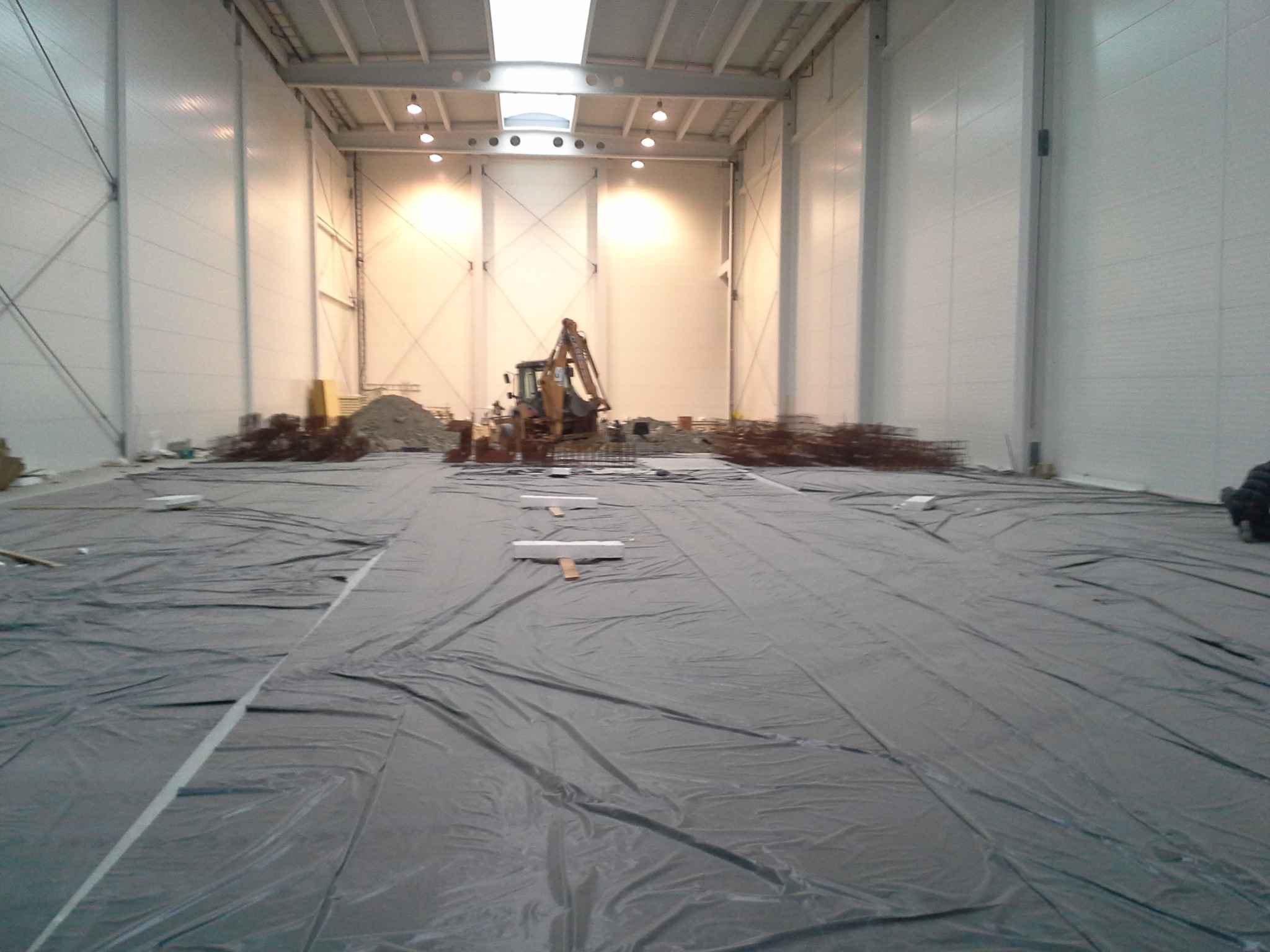 Wnętrze hali w trakcie budowy, podłoże wyłożone folią, w tle koparka i materiały budowlane.