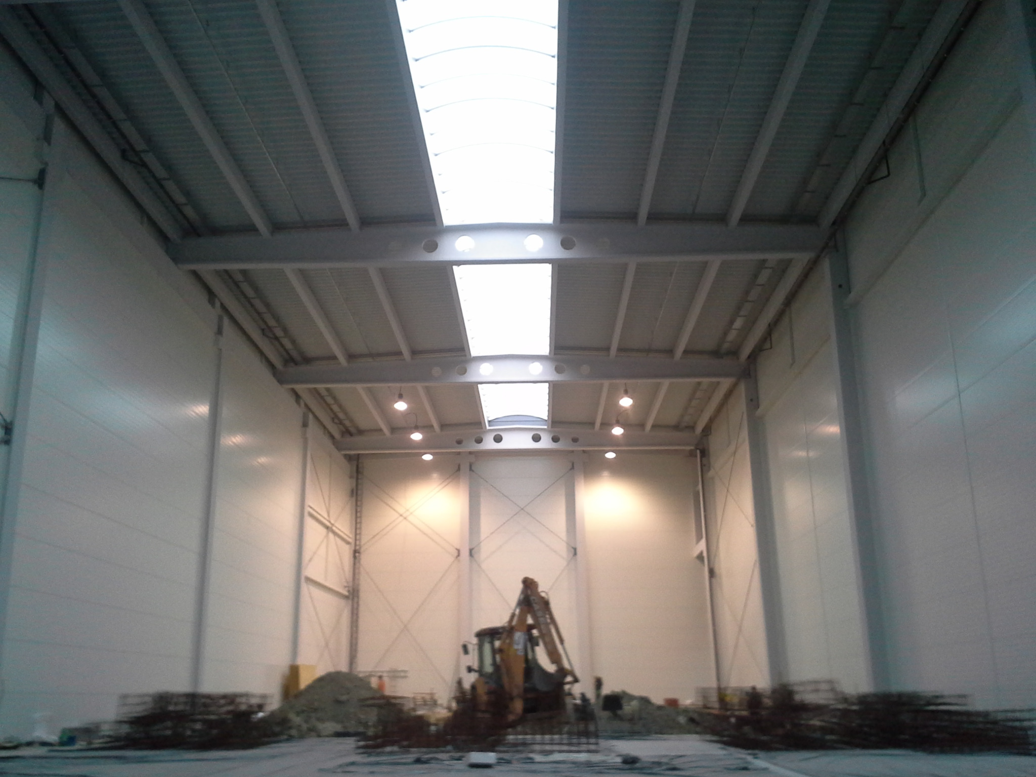 Wnętrze hali w trakcie budowy, widoczny szkielet konstrukcji, świetliki w dachu oraz koparka i materiały budowlane na posadzce.