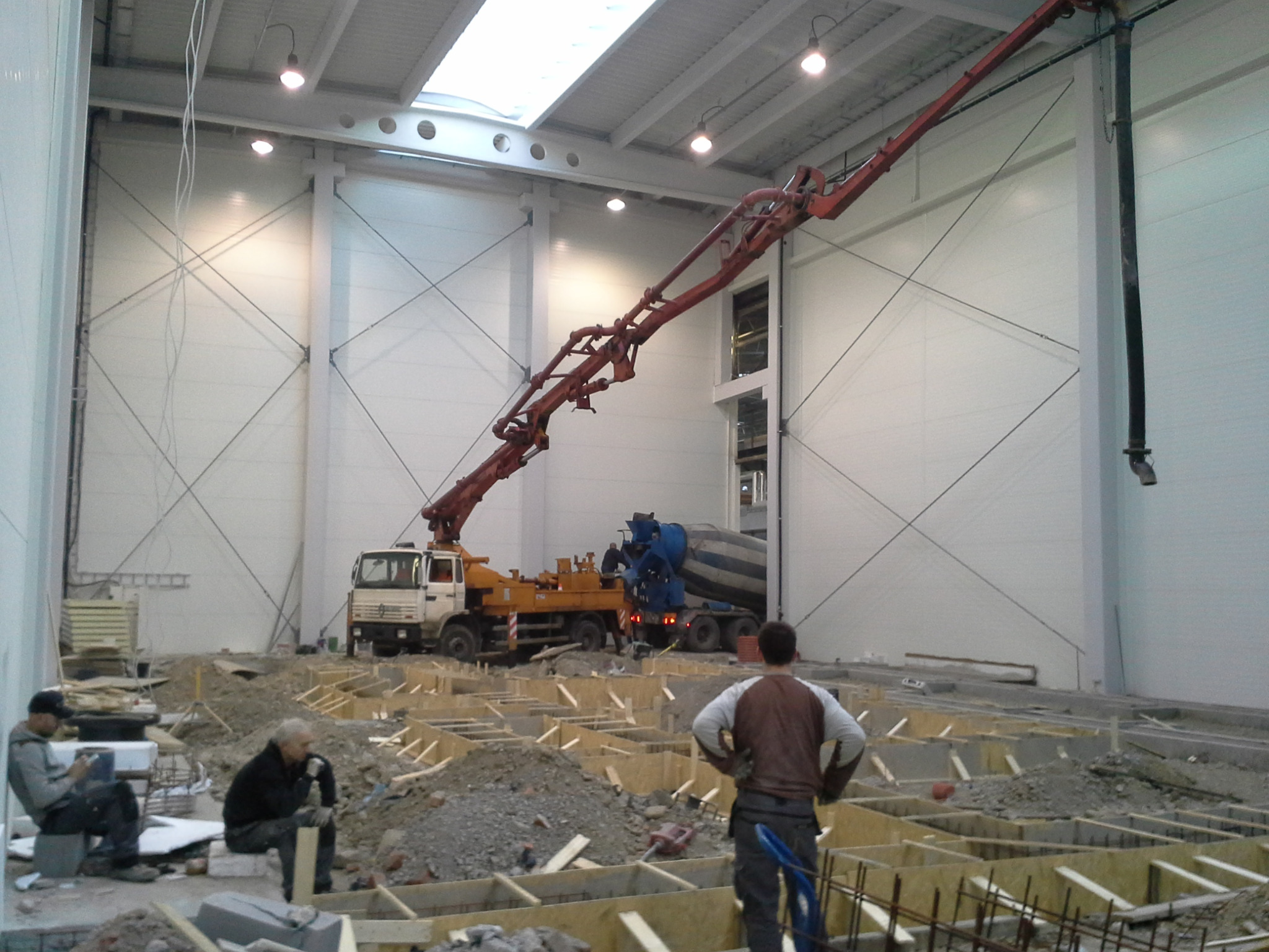 Wnętrze hali w trakcie budowy: szalunki fundamentowe, betonowóz z pompą do betonu i pracownicy w tle.