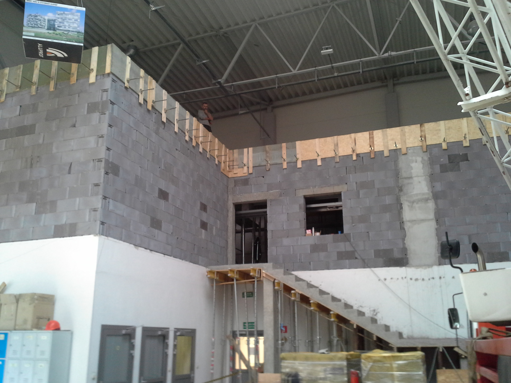 Wnętrze budowanej hali z szarych bloczków, widoczne otwory okienne i drzwiowe, drewniane elementy szalunkowe, częściowo wymurowane ściany, konstrukcja schodów tymczasowych, pracownik na górze...