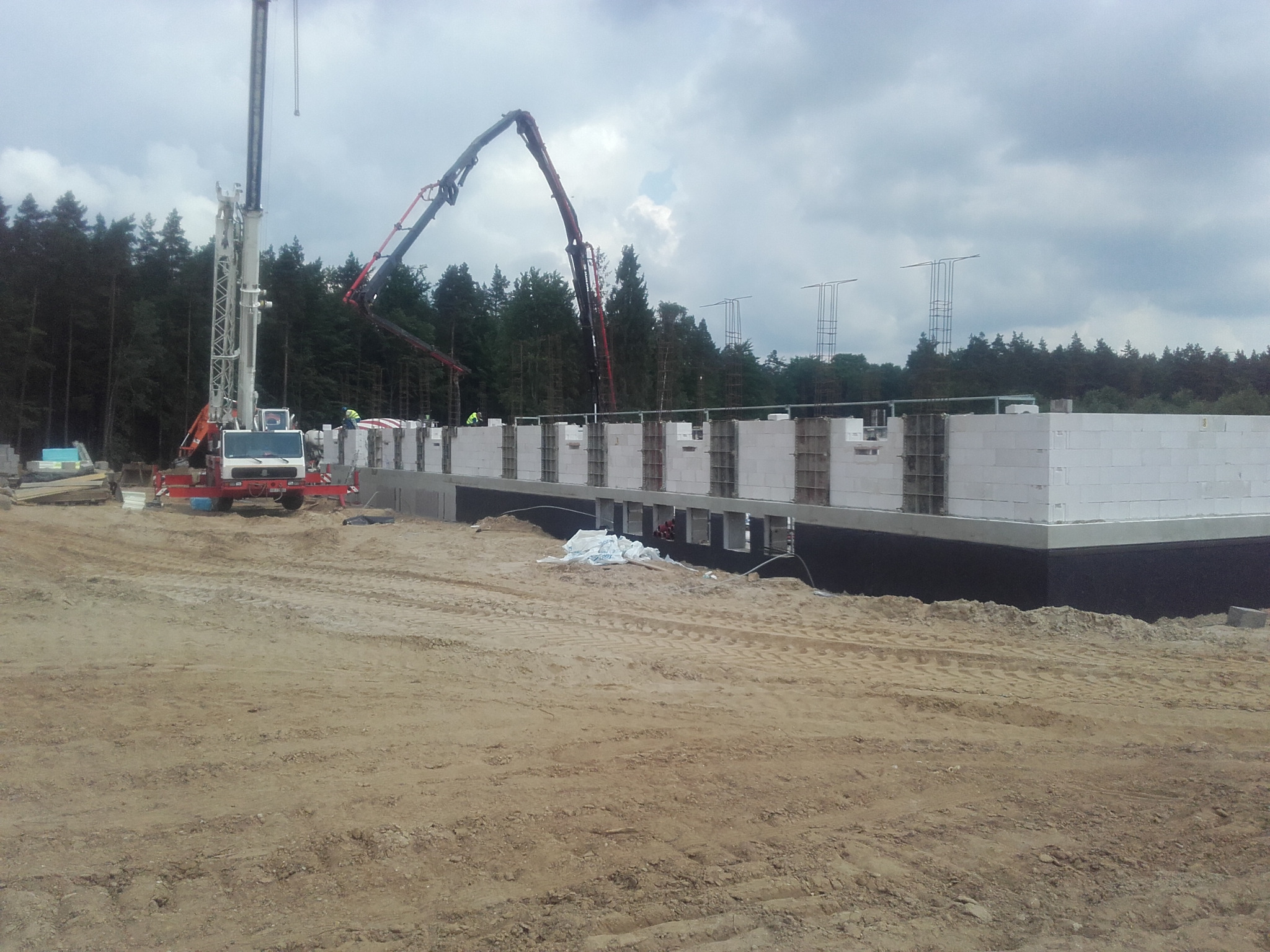 Fundamenty domu z białych bloczków, izolowane czarną folią, zbrojenia pionowe, dźwig i pompa do betonu w tle, teren budowy z piaskiem i lasem w oddali.