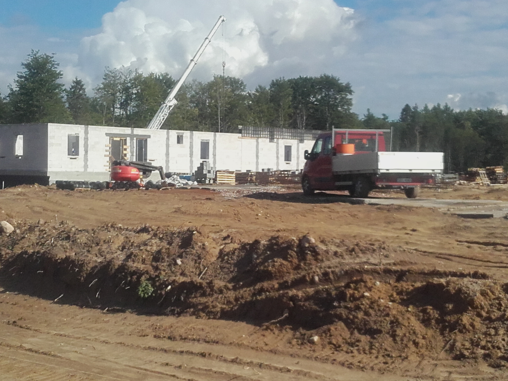 Fundamenty domu z betonu komórkowego w trakcie budowy, widoczny mini-koparka, podnośnik i ciężarówka na placu budowy.